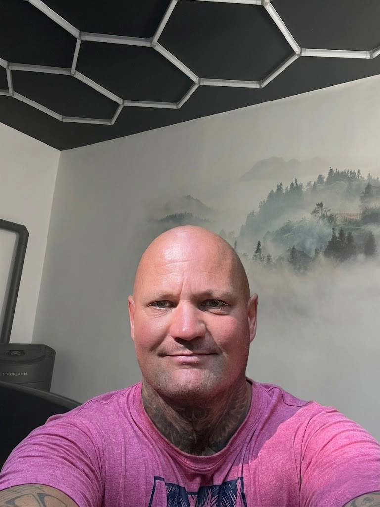 Muskulöser Mann mit Glatze und Tattoos im pink T-Shirt, Selfie im modernen Raum mit Hexagon-Deckendesign und Bergwandbild