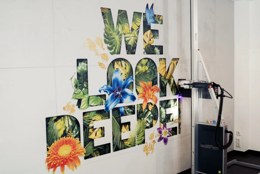 WallPen Wanddrucker druckt farbenfrohen Schriftzug WE LOOK DEEPER mit tropischen Blumen und Blättern direkt auf die Wand
